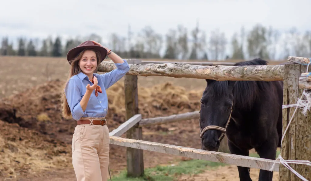 A girl on cowboy clothes stands on a horse farm near wooden fence and a black horse