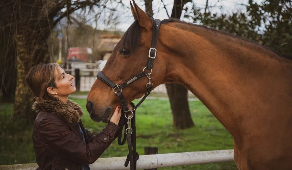 Cheerful and smiling woman dressed in a brown jacket looking at her horse at the ranch