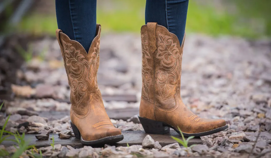 Closeup of tan leather pointed cowboy boots worn by a woman next to railroad tracks
