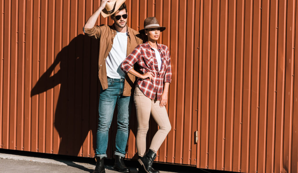 Cowboy and cowgirl in casual clothing leaning on brown wall and looking away at the ranch