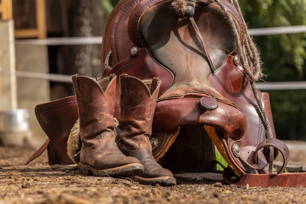 Muddy western boots and a cowboy hat in front of a western saddle 