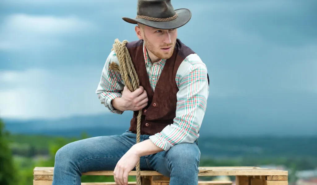Portrait of a cowboy  sitting outdoor holding his lasso rope and wearing vest 