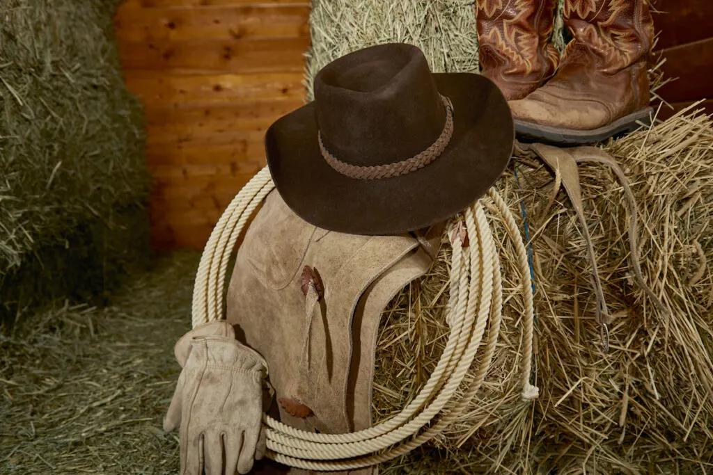 Western cowboy hat with cowboy boots, leather gloves, leather chaps and a ropers rope on hay in a barn