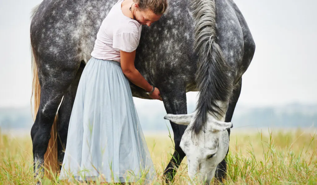 Woman in skirt with arms around dapple grey horse in field ee220401