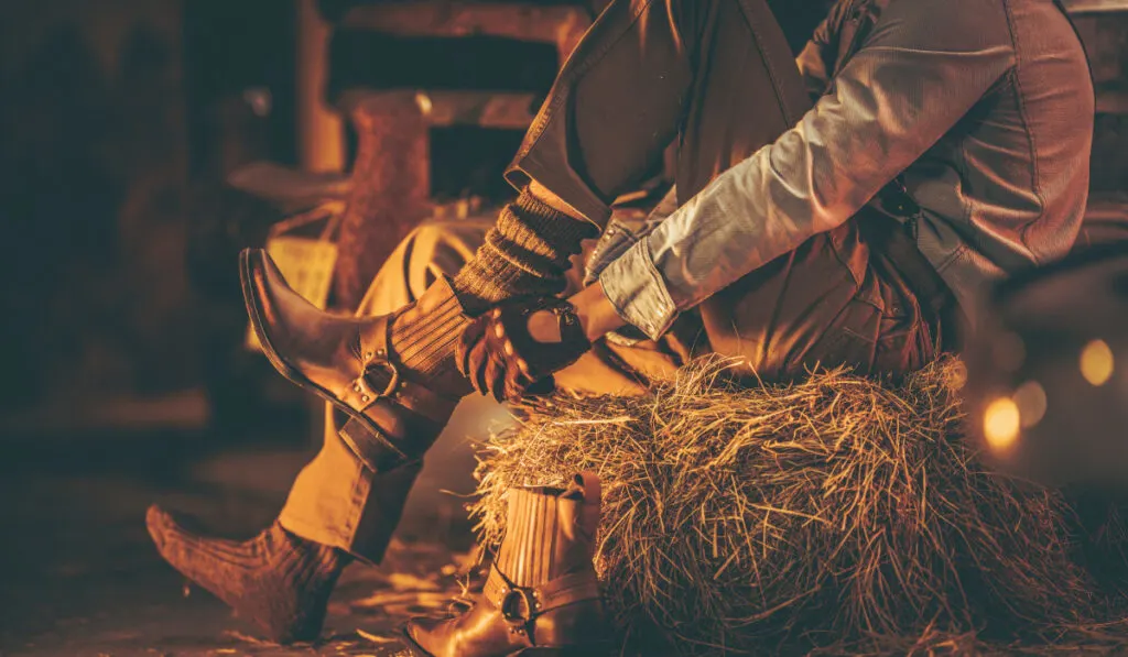 Cowboy in the Barn Wearing Boots While Seating on the Pile of Hay. 