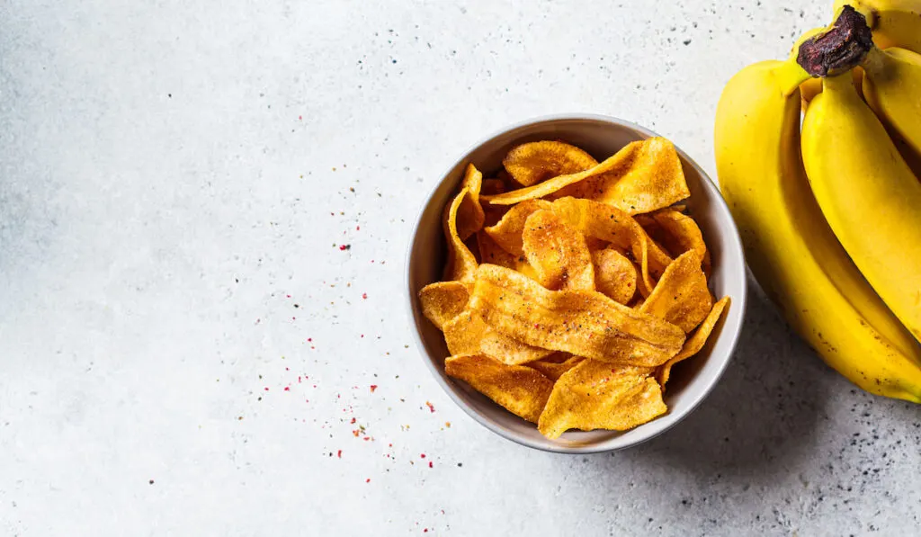 spicy banana chips in a bowl and whole yellow bananas on gray background 